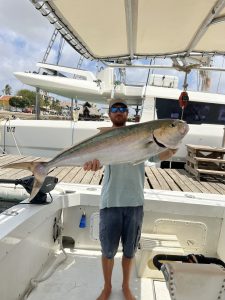 Angler in cap and sunglasses holds a large amberjack on the deck of the Fish Tales boat at the Bonaire marina dock.