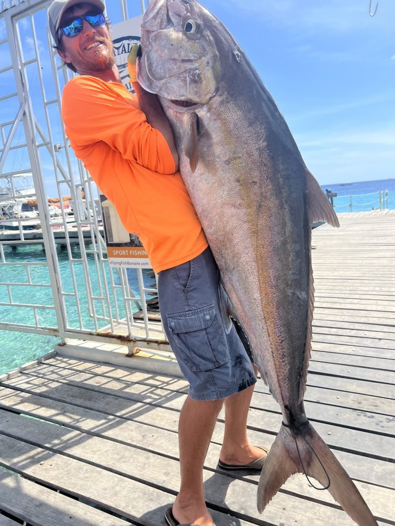 Lachende visser in oranje shirt en zonnebril houdt een enorme amberjack vast, bijna net zo lang als hijzelf, op de steiger van Bonaire.