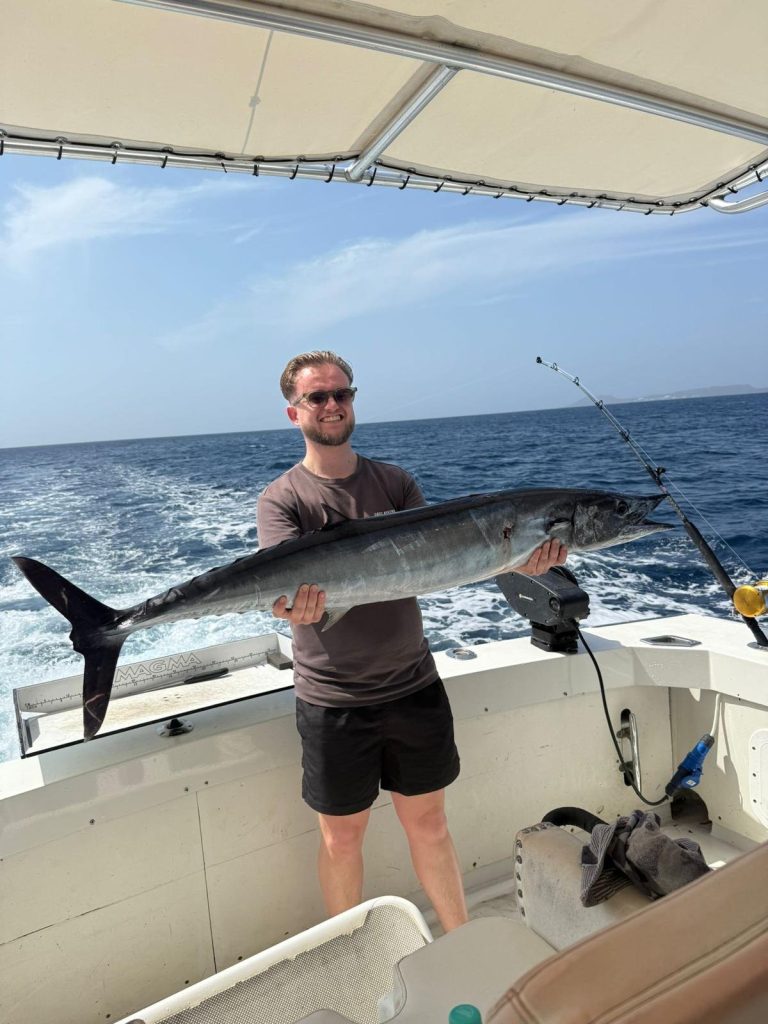 Een lachende jonge visser houdt een grote wahoo over zijn armen aan boord met de diepblauwe oceaan achter hem bij Bonaire.