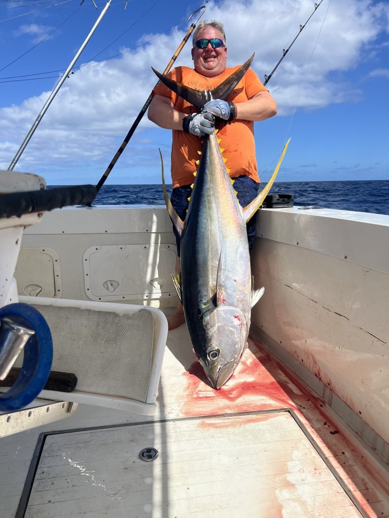 Lachende sportvisser in een oranje shirt met zonnebril tilt een zware geelvintonijn op het dek van een Fishtales sportvisser in diepblauw water.