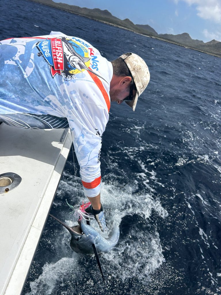 Visser in fishing-shirt met lange mouwen en pet buigt over de gunwale om boordzijds een billfish te releasen, met de kust van Bonaire op de achtergrond.