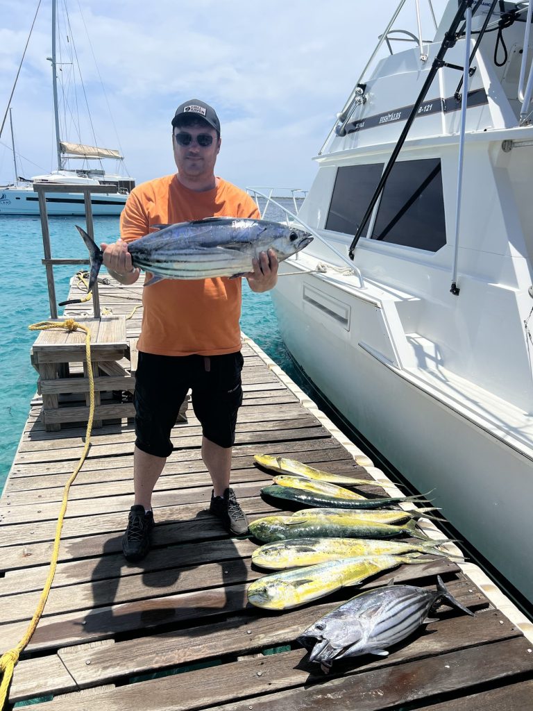 Visser in oranje shirt met een skipjacktonijn op de steiger van Nautico Marina naast de Fishtales-boot, mahi-mahi voor zijn voeten uitgelegd.