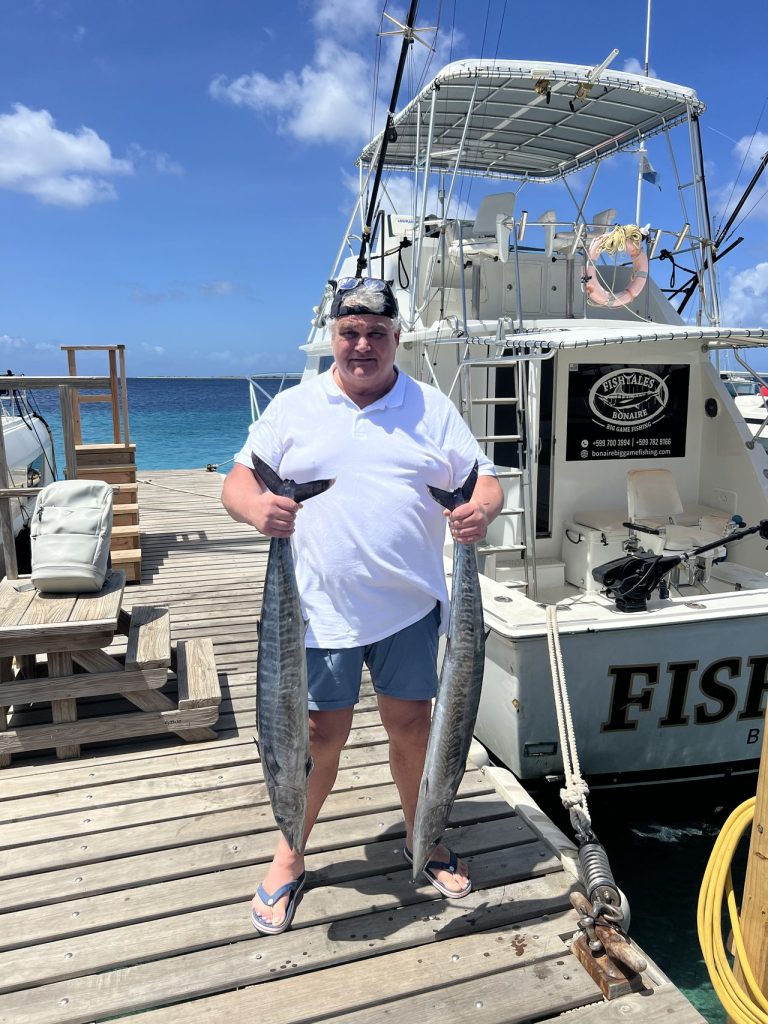 Een lachende sportvisser staat op de houten kade en houdt twee wahoo vast, een in elke hand, naast de Fishtales Bonaire sportvisser.