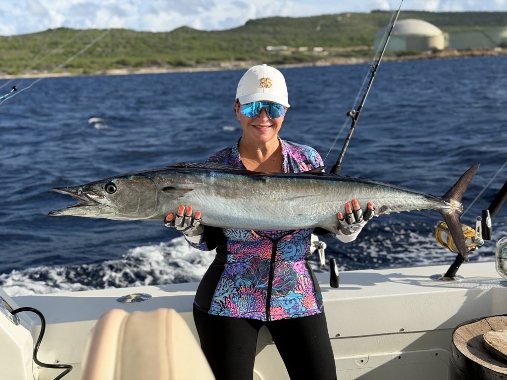 Lachende vrouwelijke visser met zonnebril en pet houdt een lange zilveren wahoo boven kobaltblauw water aan boord van een Fishtales Bonaire charter.
