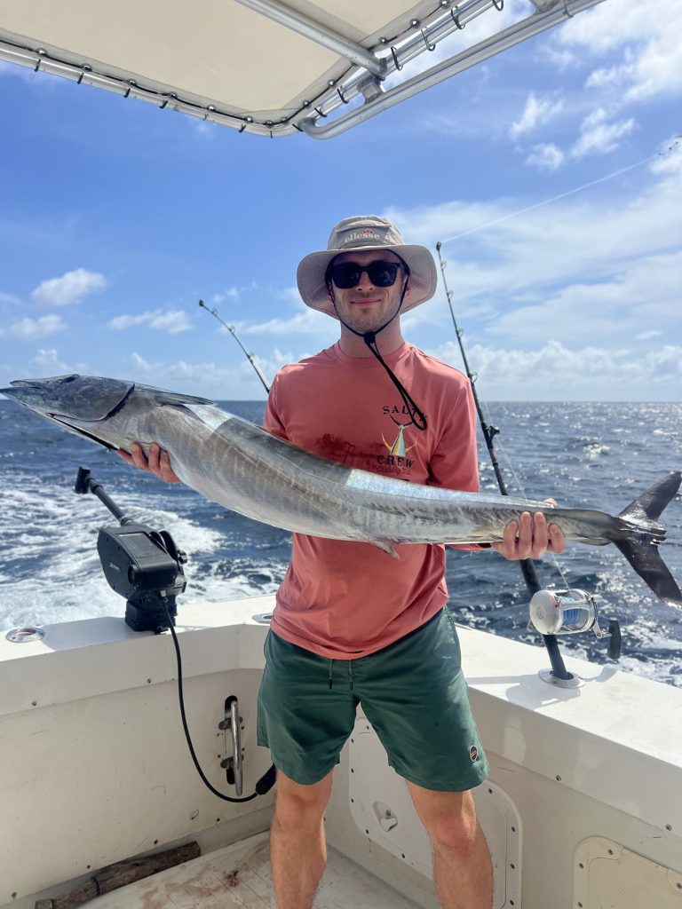 Lachende visser met zonnehoed en oranje shirt houdt een lange wahoo vast in de kuip van de Fishtales-boot onder een blauwe hemel bij Bonaire.