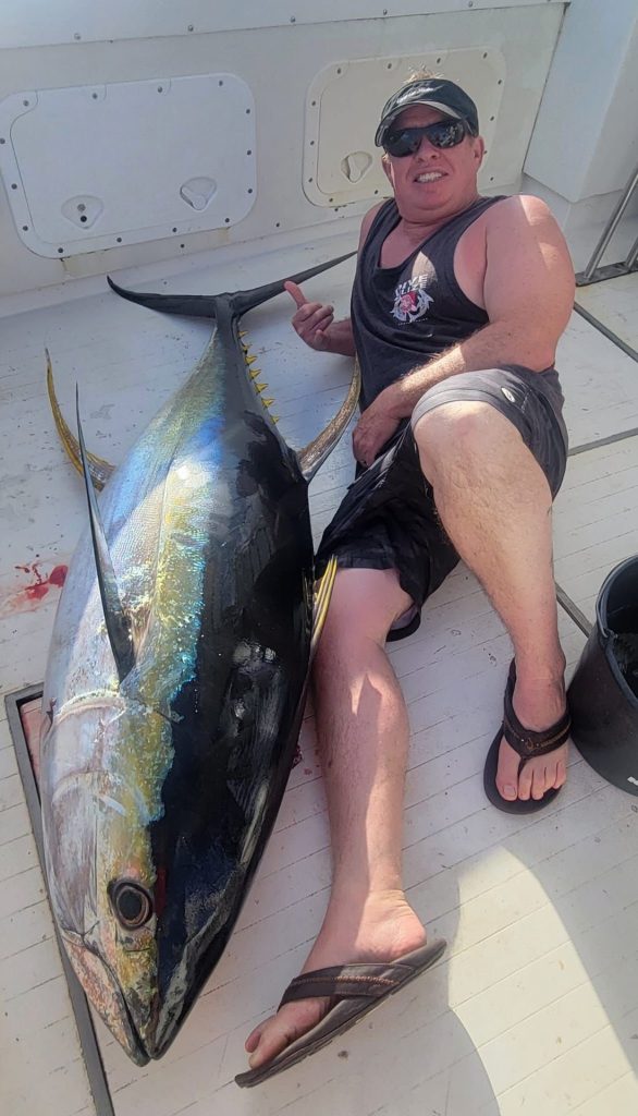 Lachende visser leunt achterover op het bootdek naast een grote geelvintonijn na een big game fishing-trip van Fishtales Bonaire.