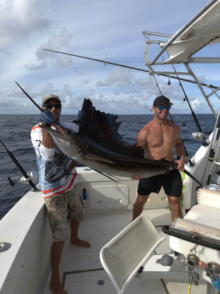 Twee vissers op de Fishtales-charterboot met een zeilvis met gespreide rugvin, open oceaan en hengels op de achtergrond voor Bonaire