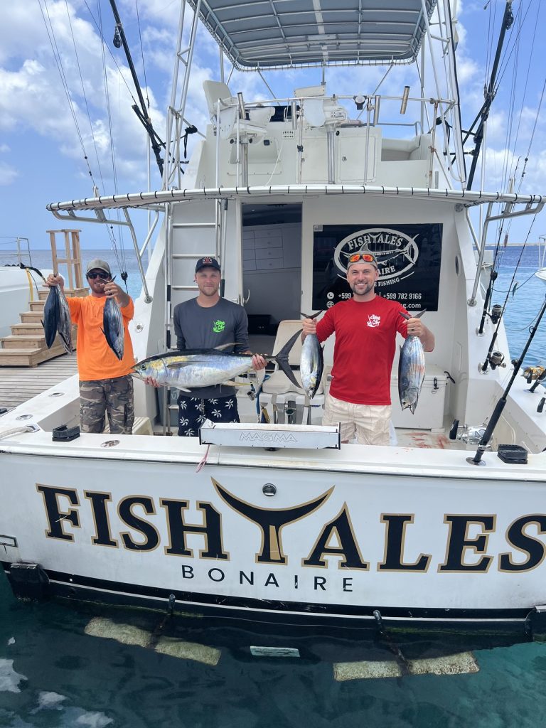 Drie sportvissers houden meerdere tonijnen vast aan het achterdek van de Fishtales Bonaire sportvisboot, met de opvallende naam FISH TALES op de spiegel.