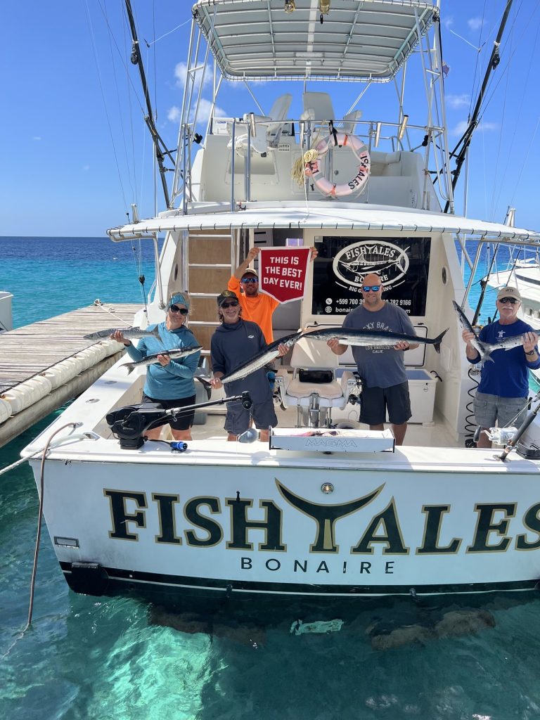 Groep vissers houdt wahoo en een Best Day Ever-bord vast aan boord van de Fishtales Bonaire-boot boven helder turquoise water.