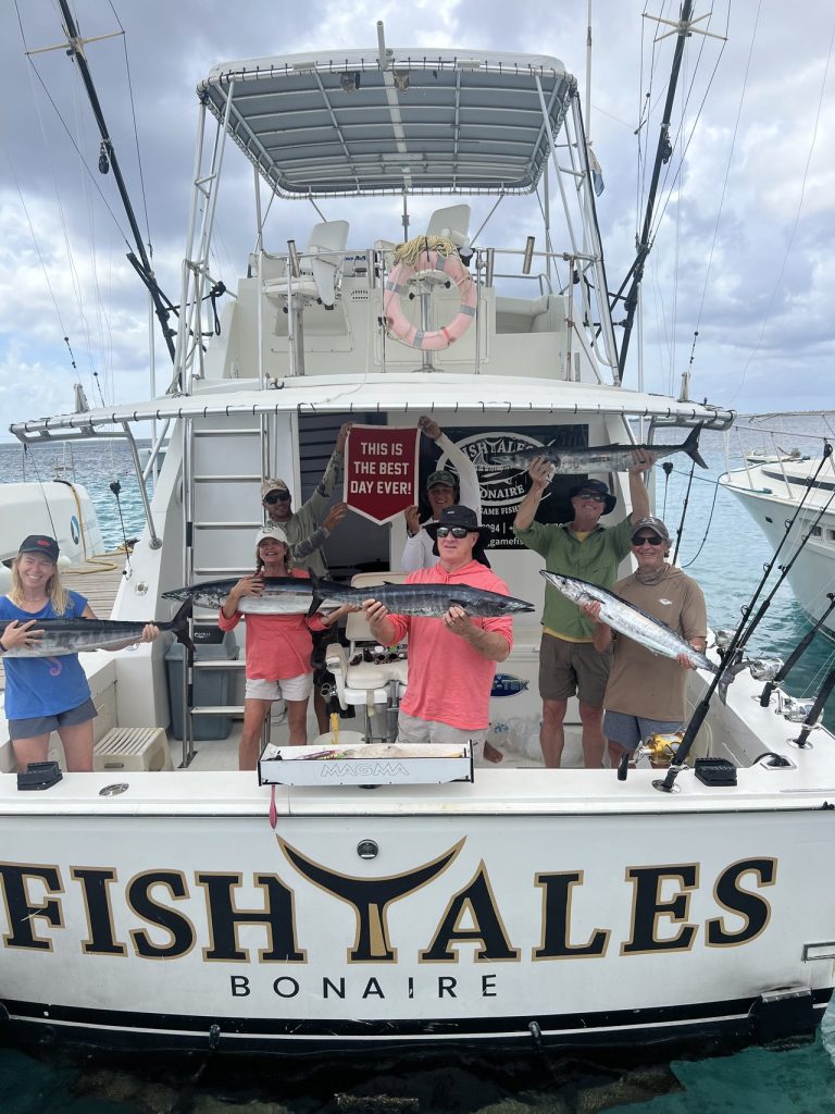 Groep vissers op de Fishtales Bonaire-boot met wahoo en een bordje This Is The Best Day Ever boven de spiegel met opschrift FISH TALES.