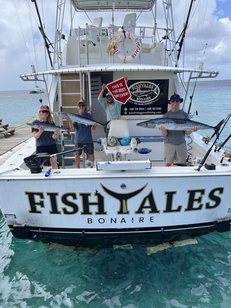 Vier vissers houden twee grote wahoo en een Best Day Ever-vlag vast op de Fish Tales Bonaire sportvisboot in turquoise water