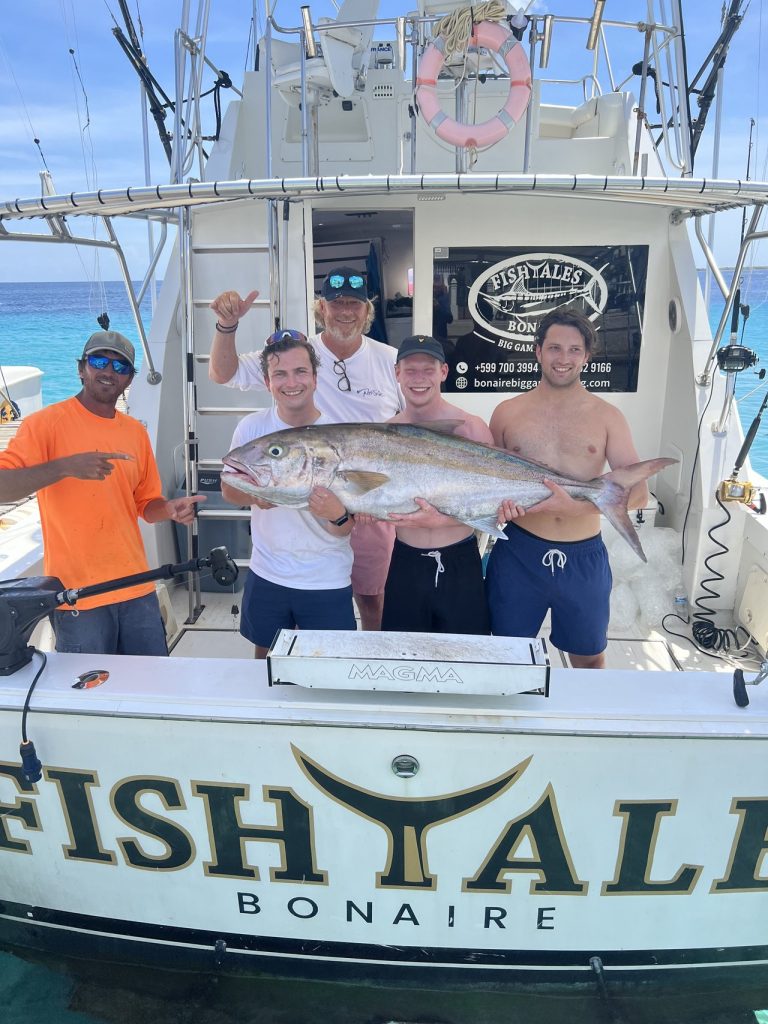 Groep van vijf lachende vissers op de spiegel van de Fish Tales Bonaire-boot met een grote amberjack boven helder blauw water.