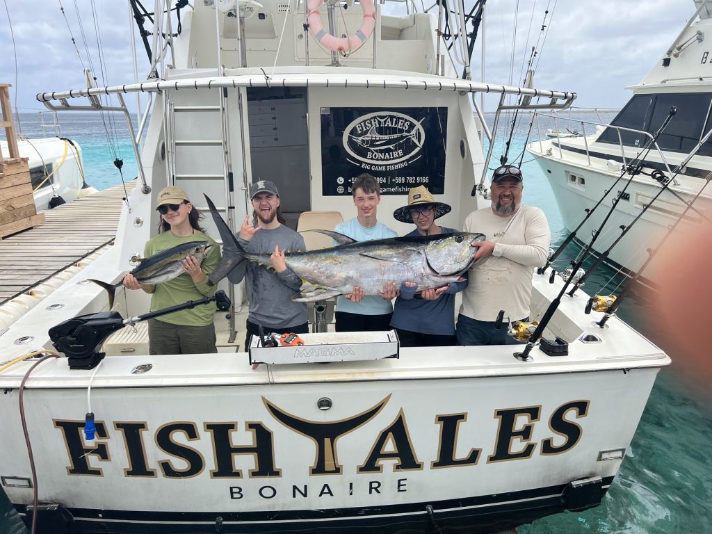 Vijf vissers poseren met een wahoo en geelvintonijn op het achterdek van de Fish Tales Bonaire sportvisboot in turquoise water.