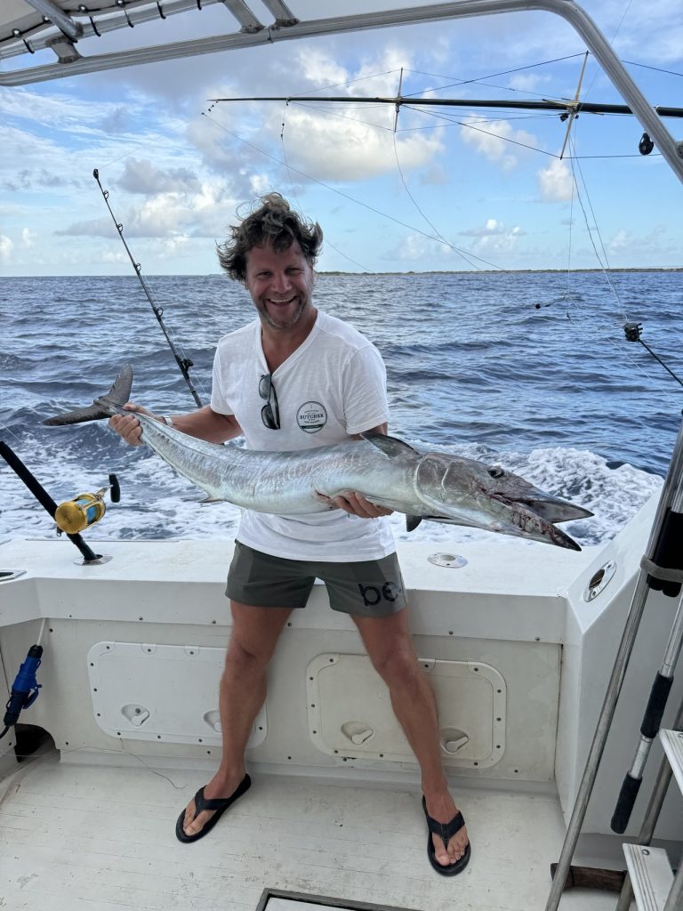 Lachende man op de boot houdt een grote gestroomlijnde big game-vis over zijn armen, met de kust van Bonaire op de achtergrond.