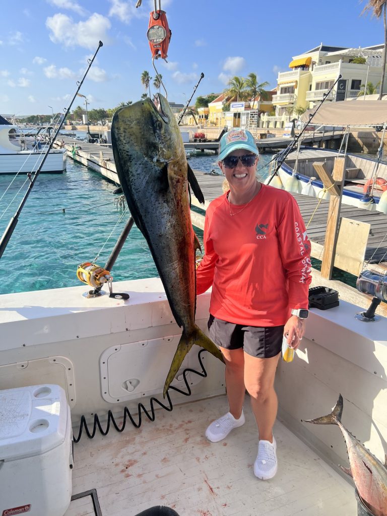 Lachende visser in een rood shirt naast een grote mahi-mahi aan de weegschaal bij de jachthaven van Kralendijk, Bonaire, met kleurrijke waterkant erachter.