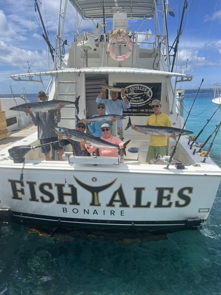 Groep van zes vissers op de Fishtales Bonaire-sportvisser met meerdere grote wahoo boven de spiegel met opschrift FISH TALES BONAIRE.