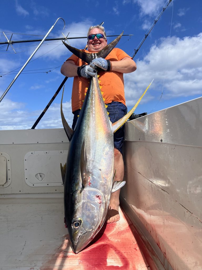 Sportvisser met zonnebril houdt een lange, zware geelvintonijn bij de staart omhoog op het dek van een sportvisboot onder een helderblauwe lucht voor de kust van Bonaire.