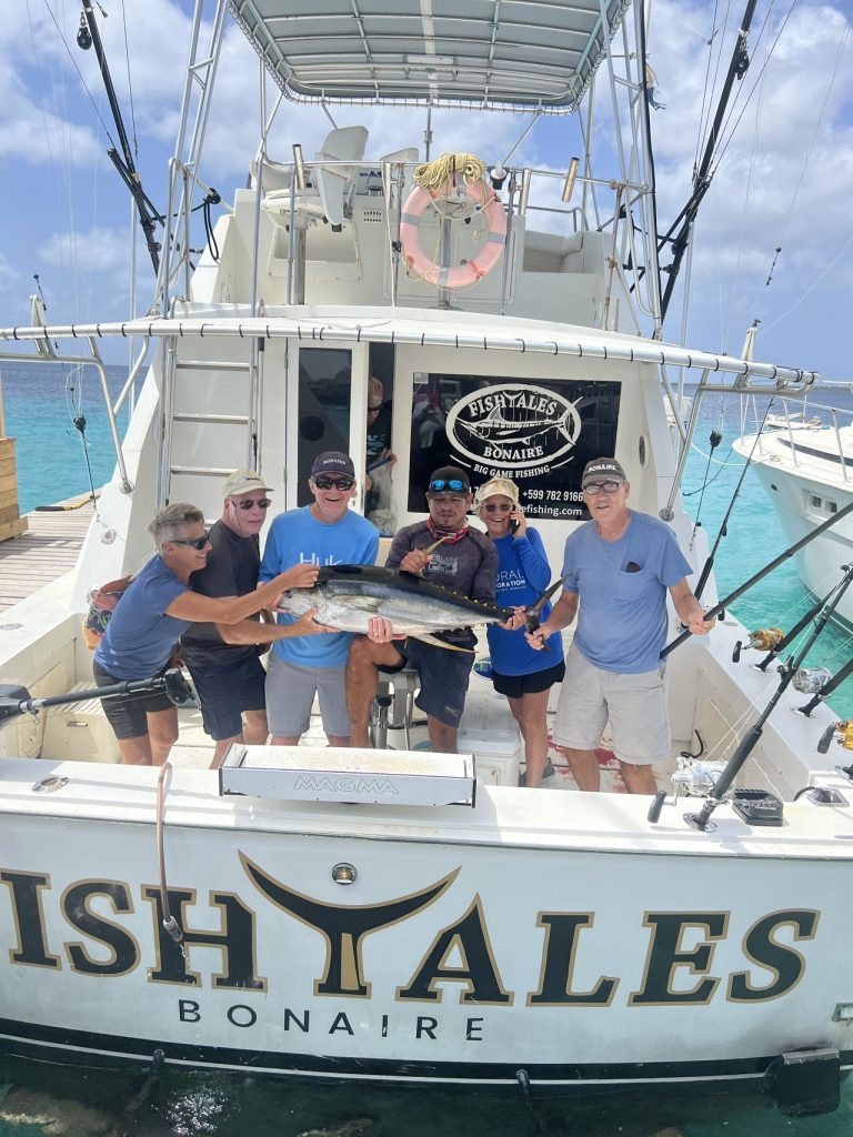 Zes sportvissers verzamelen zich aan het achterdek van de Fishtales Bonaire boot en tillen samen een grote geelvintonijn op boven turquoise water.