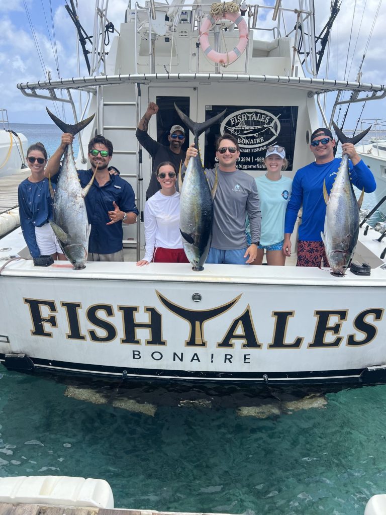 Acht vissers en bemanning houden meerdere tonijnen omhoog aan de spiegel van de sportvisser Fish Tales Bonaire boven helder water.