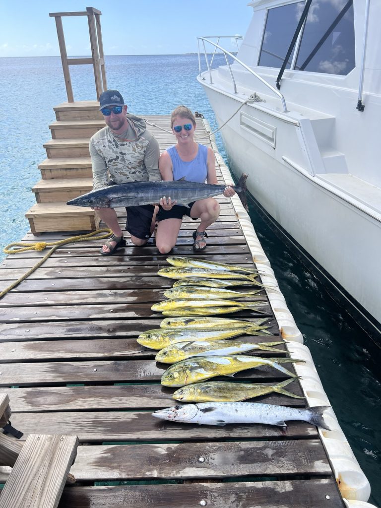 Een stel geknield op de steiger met een grote wahoo, met een rij mahi-mahi en een barracuda uitgelegd naast de charterboot.