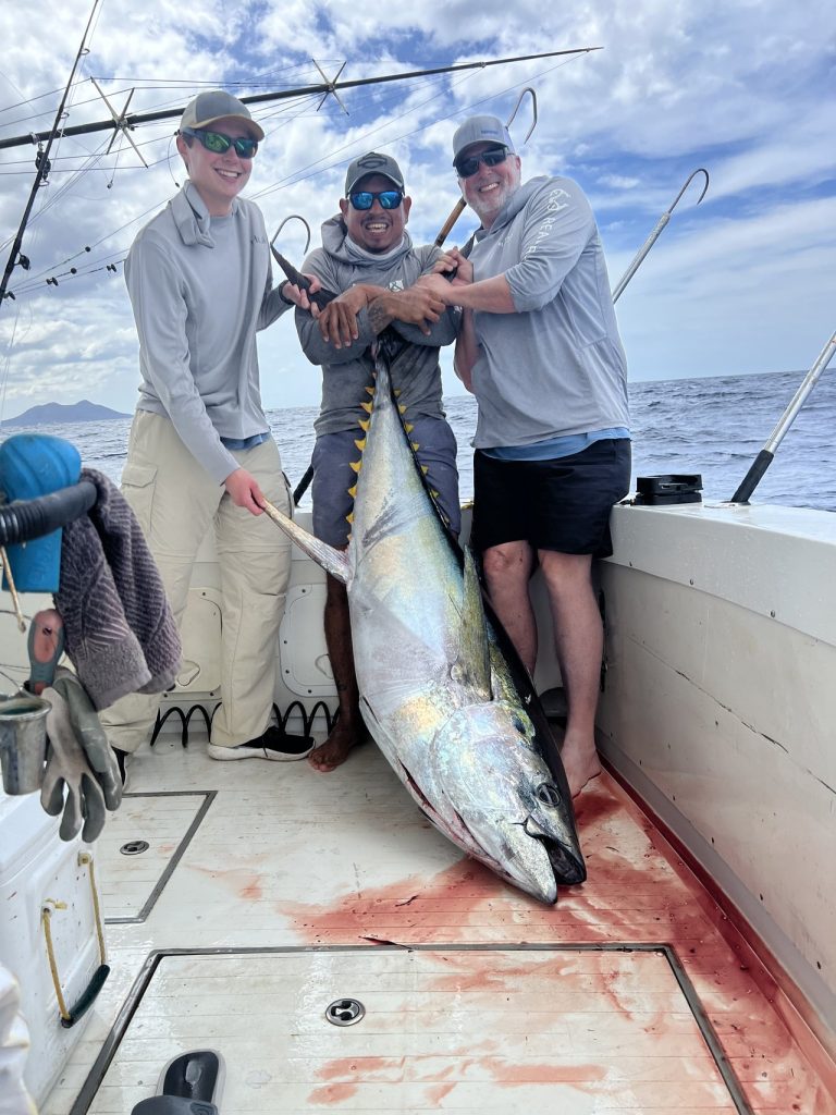 Three men, including a Fishtales crew member, grin while holding a giant yellowfin tuna on the deck of a sportfisher off the Bonaire coast.