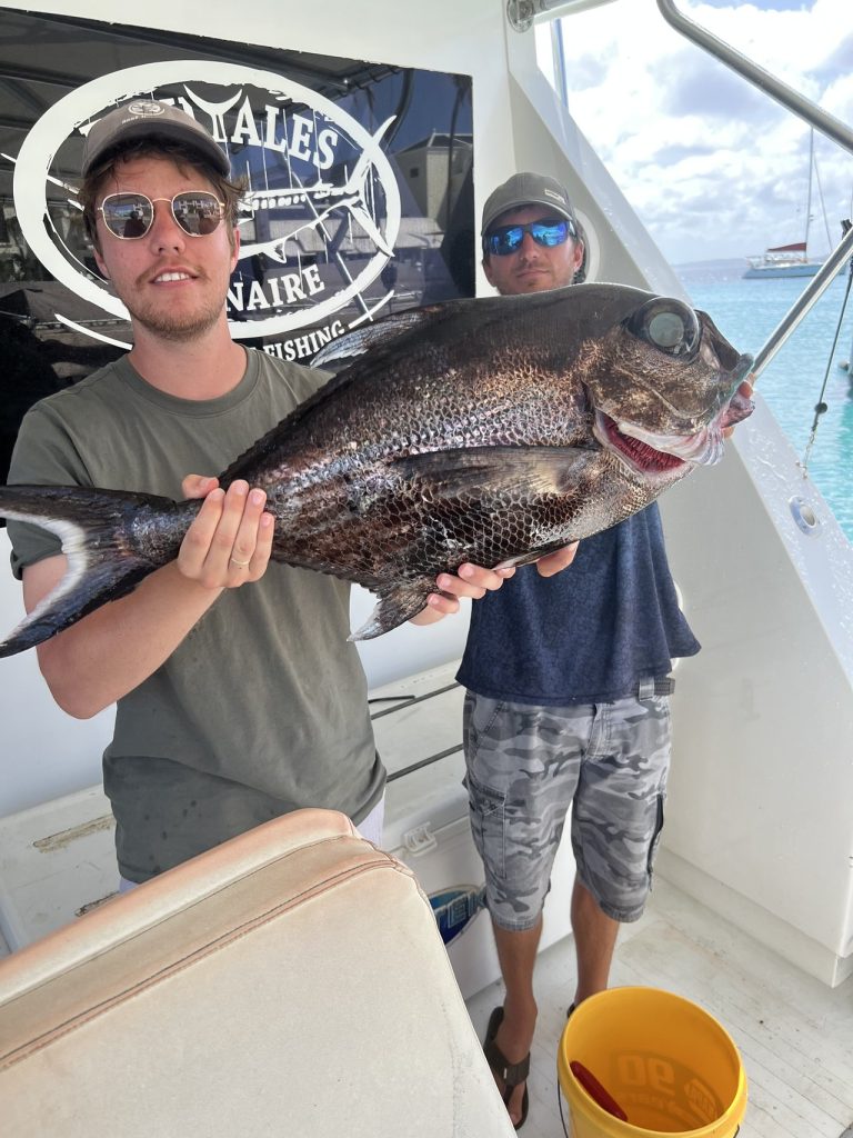 Jonge visser met zonnebril houdt een grote donkere diepwatervis vast aan boord van de Fishtales Bonaire boot met een bemanningslid erachter onder de banier.
