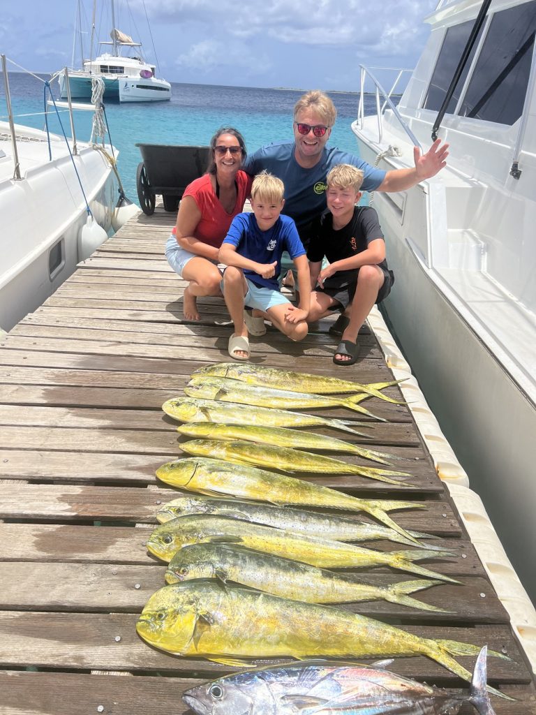 Vierkoppige familie geknield op de steiger achter een rij felle mahi-mahi en een tonijn na een Fishtales-familiecharter op Bonaire.