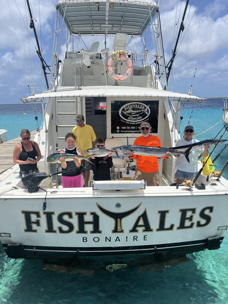 Familie van vissers aan de achtersteven van de FISH TALES Bonaire-boot met mahi-mahi en wahoo boven helder turquoise Caribisch water.