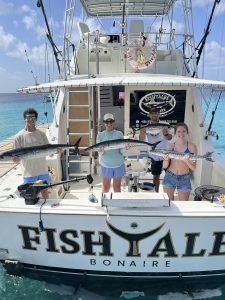 Four anglers hold wahoo and barracuda in the cockpit of the Fish Tales Bonaire sportfisher over turquoise Caribbean water.