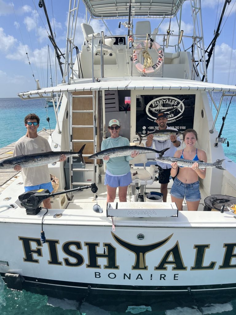 Four anglers hold wahoo and barracuda in the cockpit of the Fish Tales Bonaire sportfisher over turquoise Caribbean water.