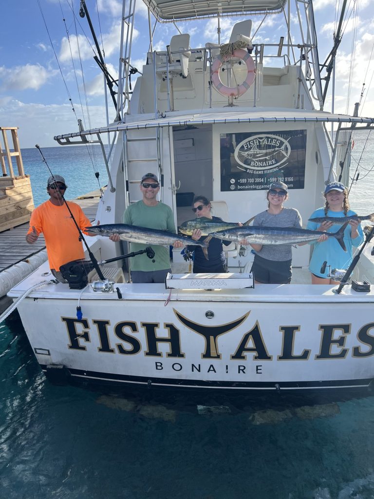 Vijf gasten houden wahoo en een mahi-mahi omhoog aan het achterdek van de Fishtales Bonaire sportvisser met de FISH TALES spiegel in beeld.