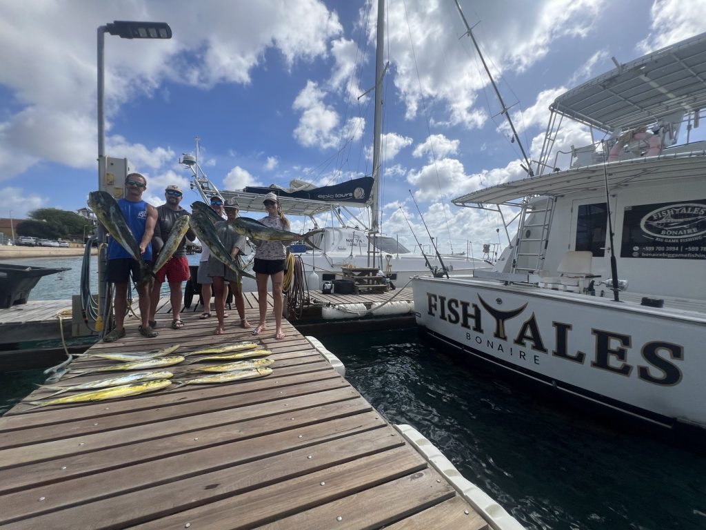 Vissers staan op de steiger van de jachthaven met mahi-mahi uitgelegd naast de Fish Tales Bonaire-sportvisser, afgemeerd in Kralendijk onder een blauwe lucht.