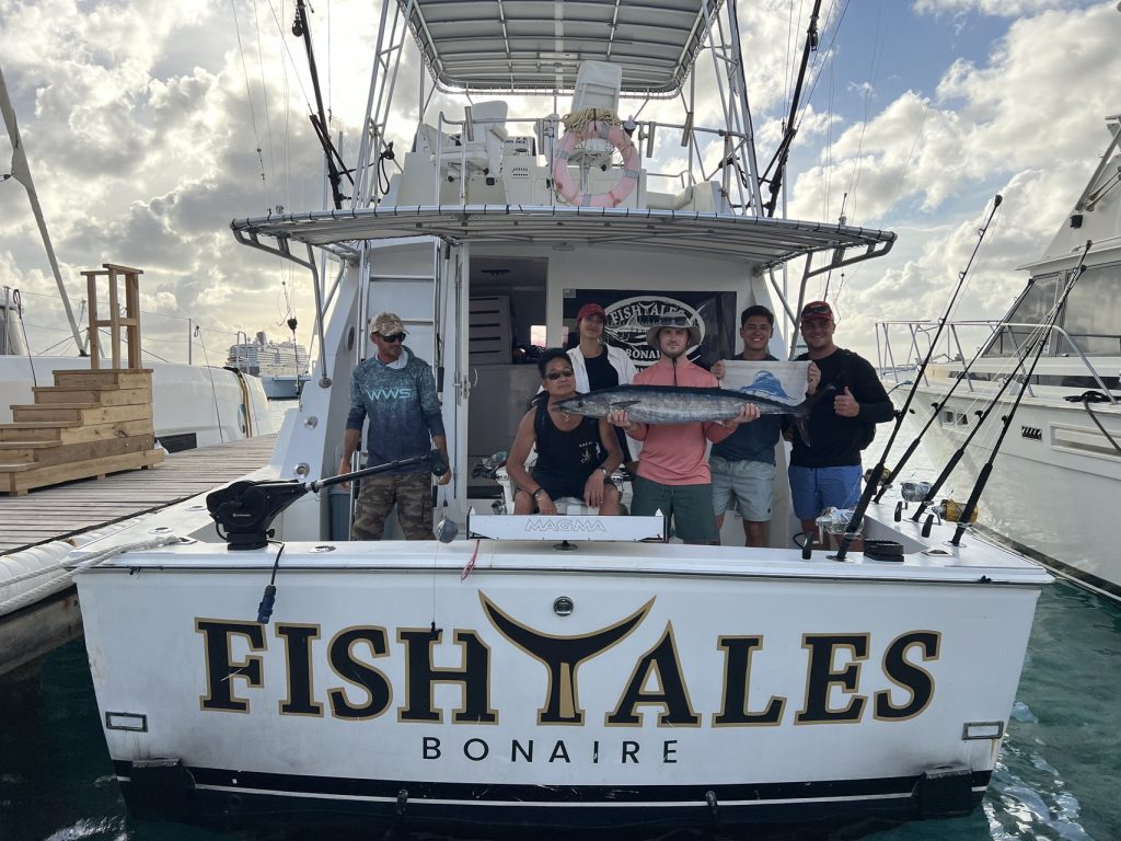 Six anglers and crew stand in the cockpit of the Fish Tales Bonaire sportfisher at the dock holding a wahoo behind the gold transom.