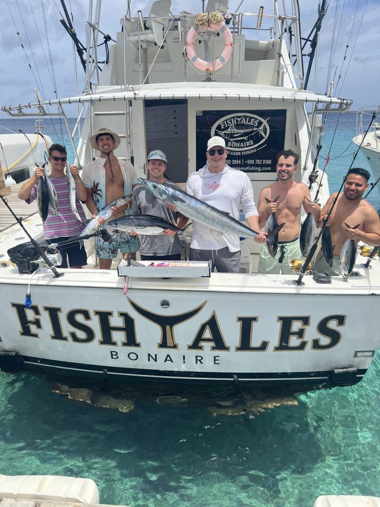 Zes vissers staan aan de spiegel van de sportvisser Fish Tales Bonaire met wahoo en tonijn boven turquoise water.