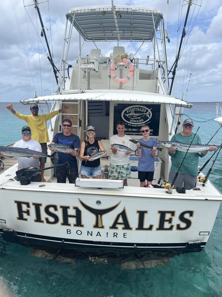 Zeven vissers staan op de Fish Tales Bonaire Bertram met wahoo en barracuda over de spiegel in turquoise water.