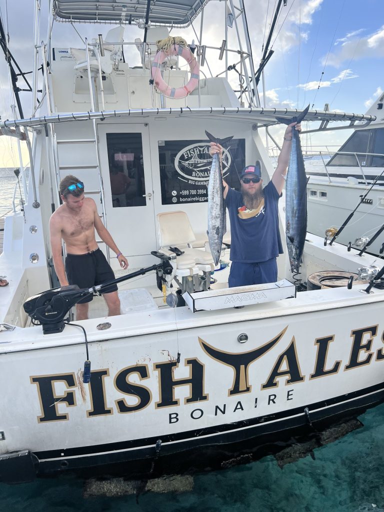 Visser houdt twee wahoo vast aan boord van de Fish Tales Bonaire-sportvisser, gouden letters op de spiegel, tweede man aan dek.