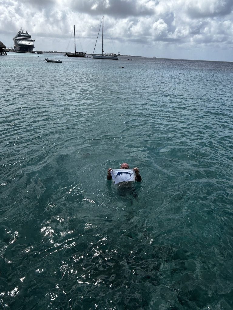 Man drijft in de turquoise haven van Kralendijk en houdt een wit Fishtales-shirt met marlijnlogo omhoog, cruiseschip en zeilboten op de achtergrond