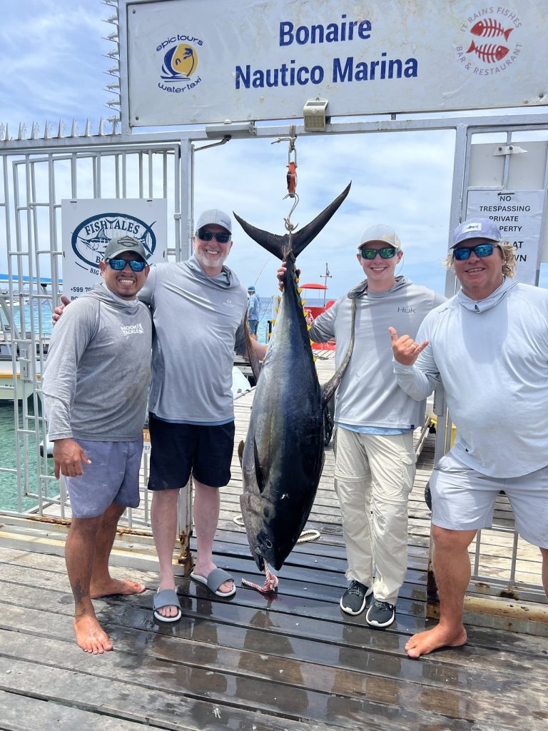 Vier sportvissers in bijpassende shirts poseren met een grote geelvintonijn die onder het bord van Bonaire Nautico Marina hangt aan de Fishtales-kade.