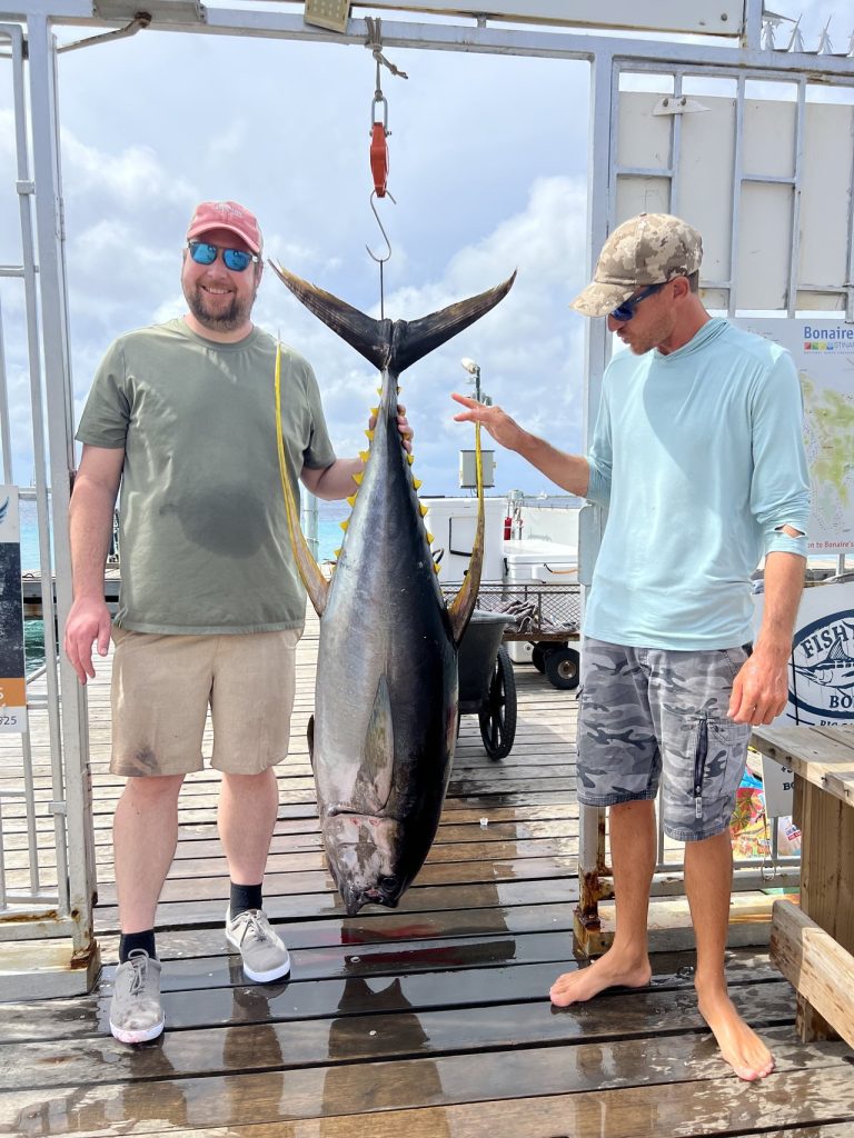 Twee mannen staan naast een reusachtige geelvintonijn die aan een weegschaal hangt op de steiger van de marina van Bonaire, met de turquoise zee erachter.
