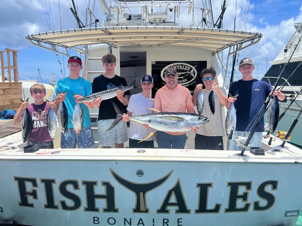 Groep van zeven jonge vissers aan de achtersteven van de Fishtales Bonaire-boot met tonijn en wahoo van hun visreis