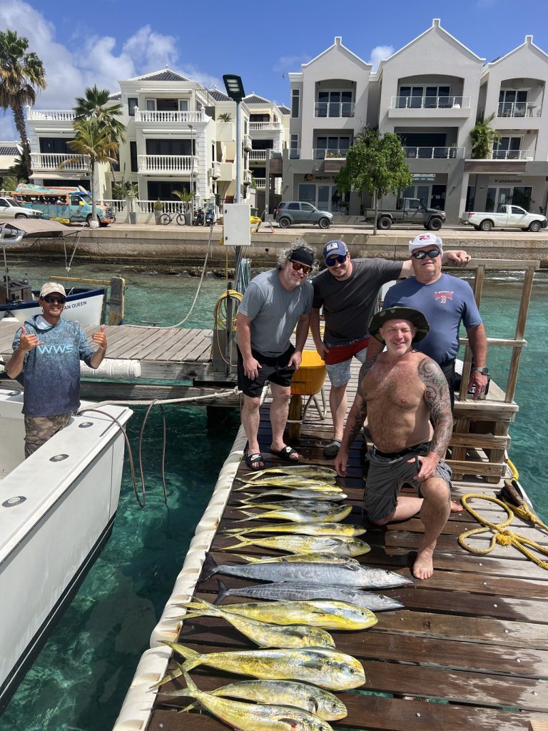 Vijf sportvissers poseren op de kade van Kralendijk met een lange rij gouden mahi-mahi, met waterfrontgebouwen achter hen op Bonaire.
