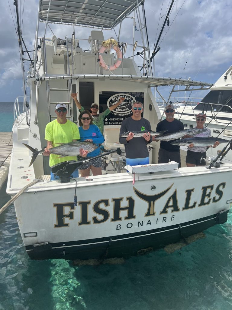Zes sportvissers houden tonijn en wahoo vast op de achterkant van de Fishtales Bonaire sportvisboot, afgemeerd in turquoise water.