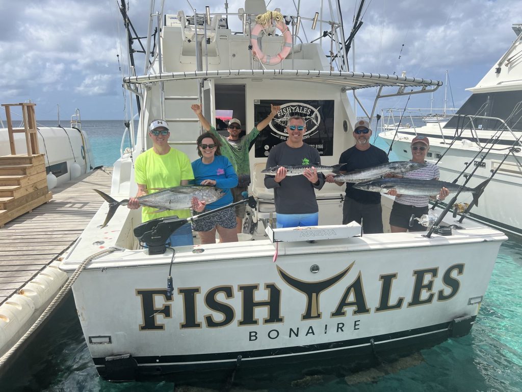 Zes gasten houden tonijn en meerdere wahoo vast over het achterdek van de Fishtales Bonaire sportvisser in helder turquoise water.