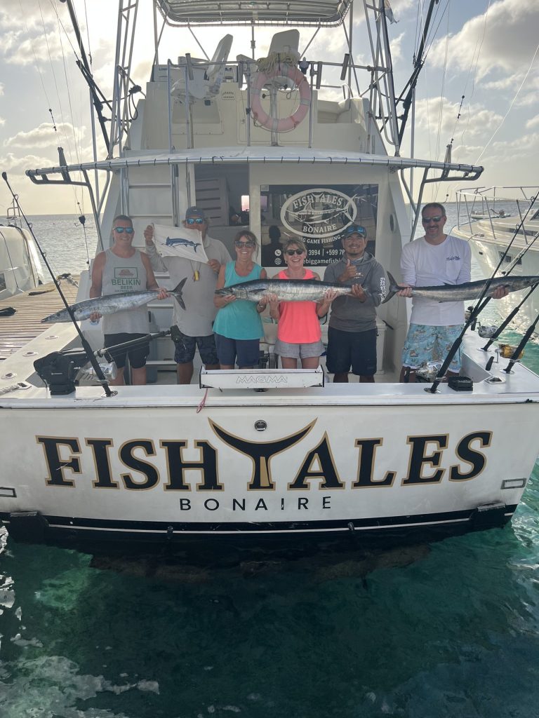 Zes gasten houden wahoo en een billfish-releasevlag vast op het achterdek van de Fishtales Bonaire sportvisser tijdens het gouden uur.