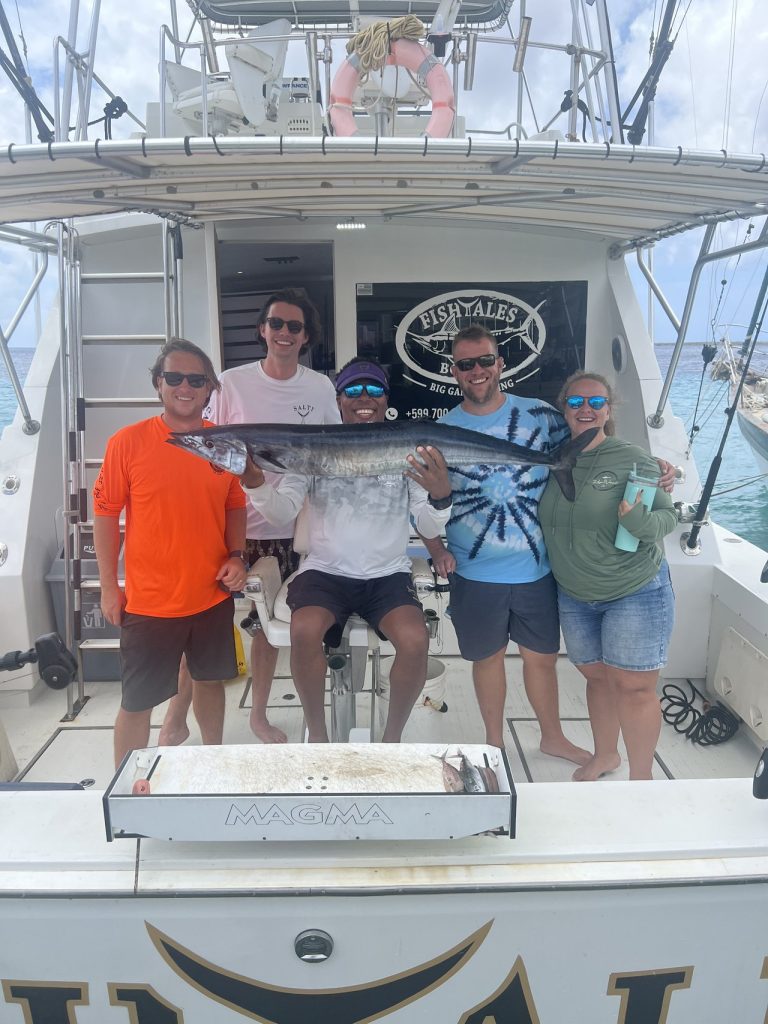 Five smiling guests on the back of the Fish Tales Bonaire sportfisher holding a long silver wahoo under the charter banner.