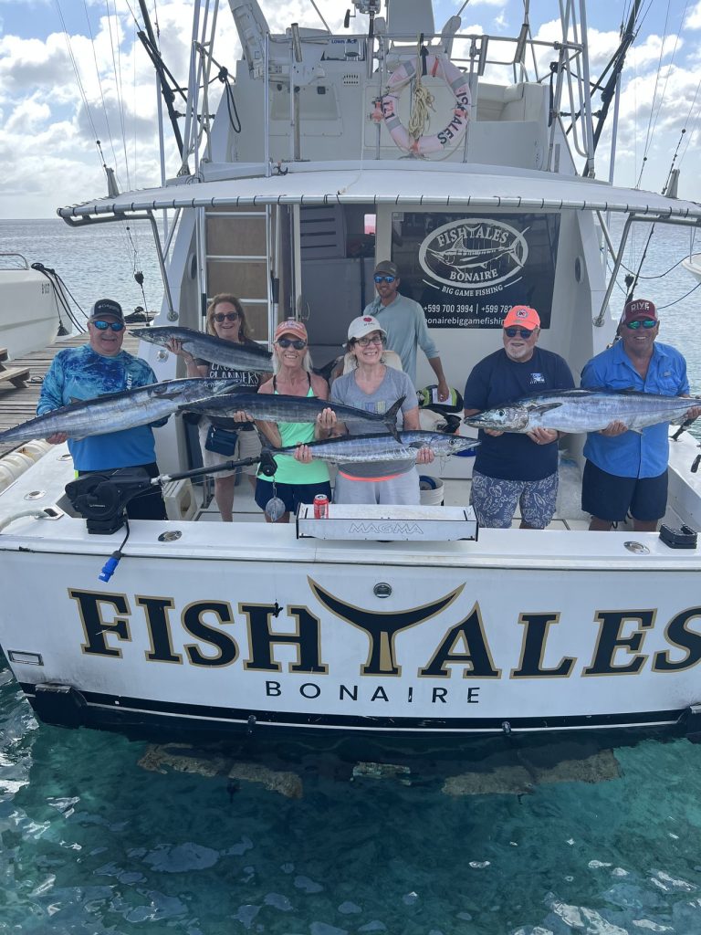 Zes vissers en bemanning houden meerdere wahoo vast over de achtersteven van de Fish Tales Bonaire charterboot in helder blauw water