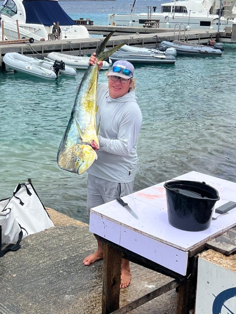 Visser houdt een kleurrijke mahi-mahi (dorado) omhoog bij het fileerstation van de haven met boten afgemeerd op de achtergrond op Bonaire.