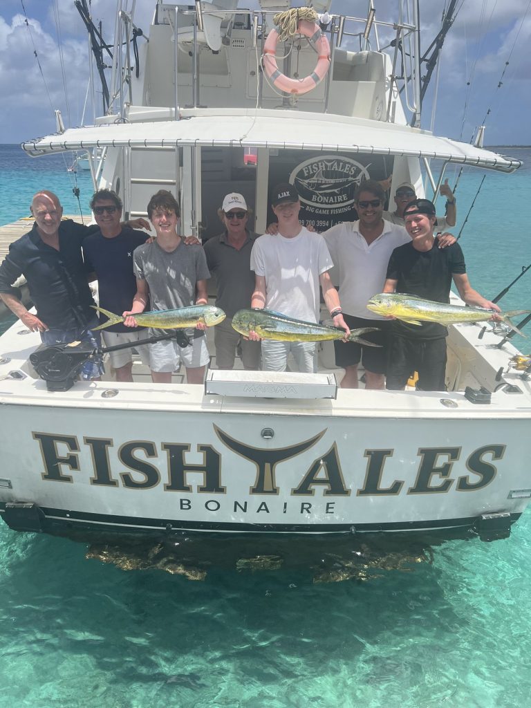 Zes vissers op de spiegel van de Fish Tales Bonaire houden drie felgouden mahi-mahi vast boven het heldere turquoise Caribische water.