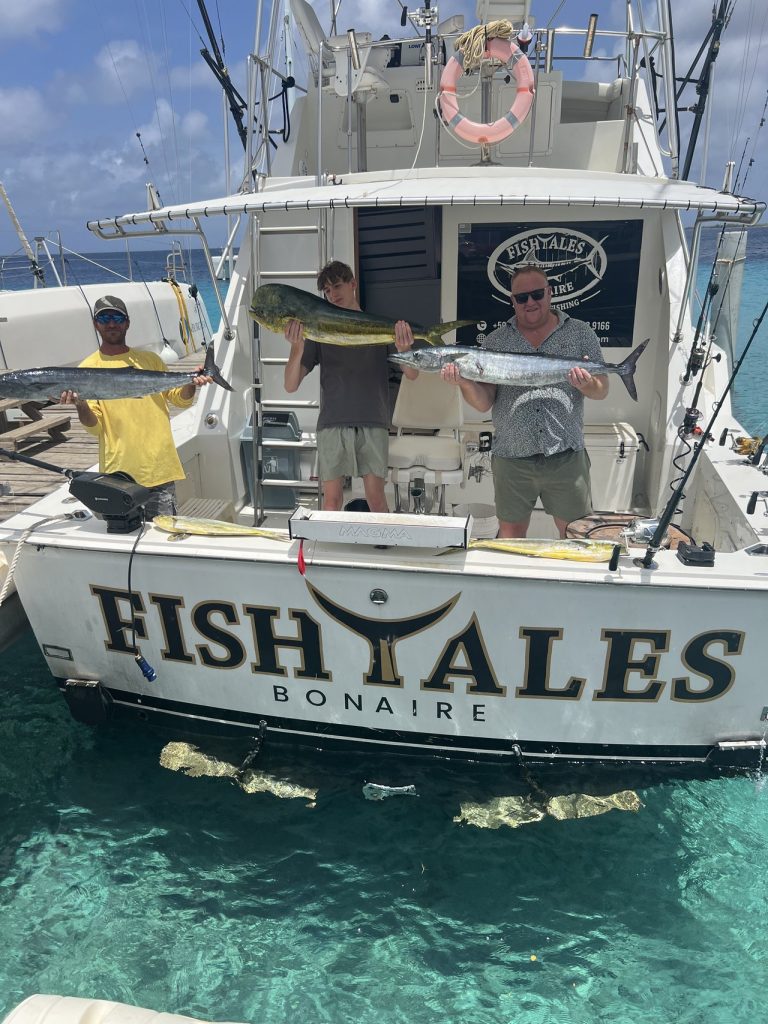 Drie vissers op de Fish Tales Bonaire boot houden een gouden mahi-mahi en wahoo vast boven helder turquoise Caribisch water.