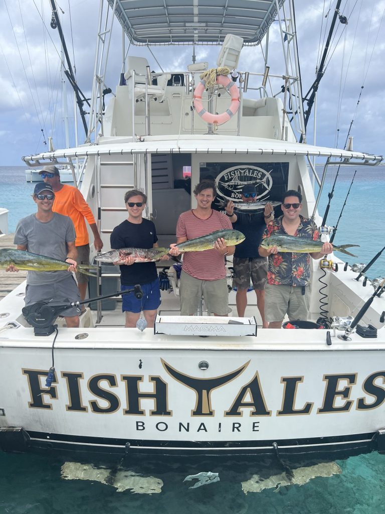 Vijf vissers en bemanning op de spiegel van de Fish Tales Bonaire houden gouden mahi-mahi en een wahoo vast boven turquoise water.